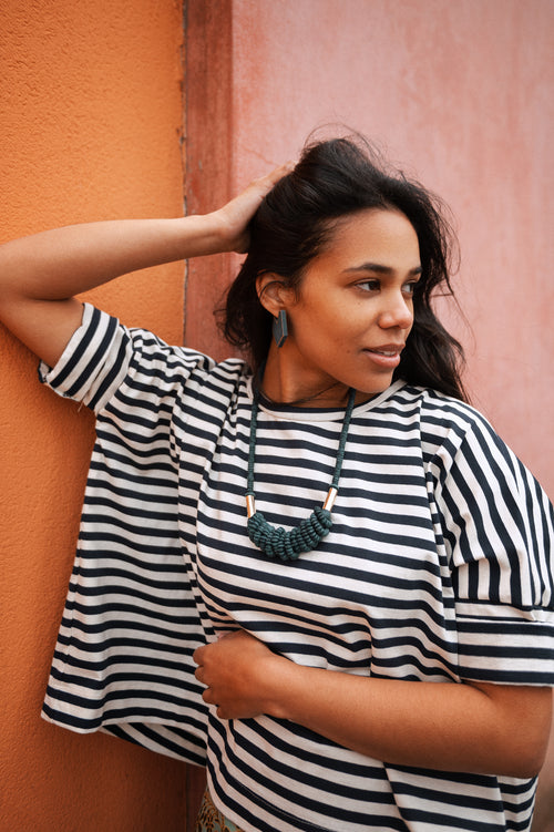 Model in front of orange pink colored wall. Model wears a white and blue striped T-shirt and petrol colored wooden necklace with geometric earrings in petrol with gold rim.